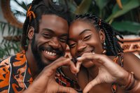 Young African couple making heart shape with hands, showing genuine joy and love while wearing traditional pattern clothing against tropical background.