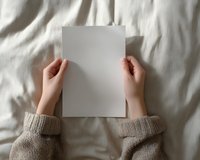 Hands holding blank white paper sheet on beige textile background with soft shadows. Cozy minimal composition with knitted sweater sleeves.