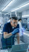 Middle-aged Caucasian male scientist in blue scrubs examining holographic molecular structure in modern laboratory with safety glasses and gloves.