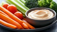 Fresh vegetable crudités platter with carrots, celery, tomatoes and broccoli served alongside creamy hummus dip in dark ceramic bowl on gray plate.
