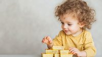 Caucasian toddler with curly hair in yellow sweater playing with wooden blocks against light gray wall background, showing curiosity and development through play.