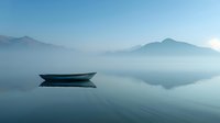 Tranquil wooden boat floating on misty lake at dawn, mountains silhouetted against blue sky, calm water surface creating mirror reflection. For travel and meditation concepts.