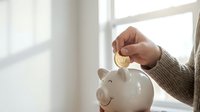 Hand inserting golden coin into white ceramic piggy bank by bright window for savings and financial planning goals.