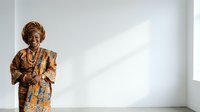 Senior African woman wearing traditional gold and blue patterned clothing with beaded necklaces, standing confidently in bright studio setting with white wall background.
