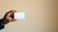 Close-up of male hand with dark skin tone holding blank white business card against neutral beige background for branding and corporate messaging.