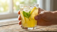 Fresh lemonade with mint and lemon slice in glass held by hand on wooden table near bright window for summer refreshment and healthy hydration concept.