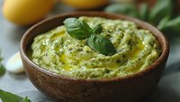Creamy basil pesto sauce in wooden bowl with olive oil drizzle, garnished with fresh basil leaf, blurred lemons in background.