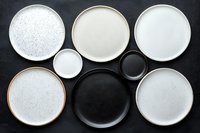 Modern ceramic dinnerware collection with neutral plates and bowls in white, black, and beige tones arranged on dark surface for kitchen and dining decor.