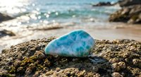 Turquoise gemstone resting on rocky shore with sparkling ocean waves and sandy beach in soft focus background creating peaceful coastal scene.