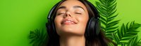 Young woman with closed eyes wearing black headphones enjoying music against vibrant green background with tropical palm leaves creating relaxing atmosphere.