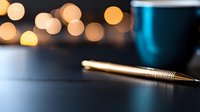 Golden pen on dark surface with blurred bokeh lights in background and partial view of blue coffee cup, creating cozy atmosphere for writing or working.