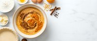 Homemade pumpkin pie filling in white bowl with baking ingredients flour eggs butter cinnamon spices on marble counter top view for autumn dessert recipes.
