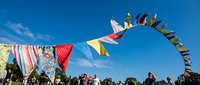 Colorful bunting flags flutter against bright blue sky at outdoor festival with crowd of people celebrating below on sunny day.