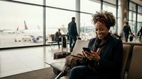 Young African American businesswoman using smartphone while waiting at airport terminal with luggage, smiling at mobile device during travel delay or layover.