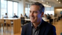Middle-aged Caucasian male professional in business casual attire smiling in modern office environment with colleagues working at desks in background. Corporate workplace setting.