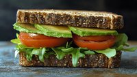 Fresh sandwich with whole grain bread, layered with ripe tomato slices, creamy avocado, and crisp arugula leaves on rustic background. Perfect for food blogs and menus.
