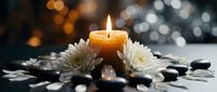 Burning candle with white chrysanthemum flowers and black spa stones creating peaceful meditation atmosphere with soft bokeh lights background.