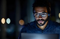 African American male customer service representative wearing glasses and headset works at night, illuminated by computer screen in dark office setting.