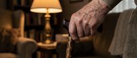Senior man's weathered hand gripping wooden walking cane in warm living room with lamp creating cozy atmosphere for elderly care and mobility support.