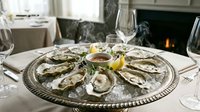 Fresh raw oysters on half shell served on silver platter with lemon wedges and mignonette sauce in elegant restaurant setting with wine glasses and fireplace.