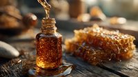 Golden honey dripping into small glass bottle beside fresh honeycomb on rustic wooden surface, warm sunlight highlighting amber tones.