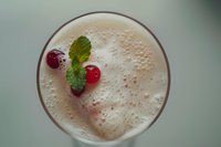 Creamy milkshake topped with fresh red cranberries and mint leaves, viewed from above. Frothy drink surface shows delicate bubbles against soft green background.