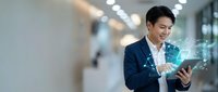 Young Asian businessman in navy suit using digital tablet with holographic data visualization in modern office corridor for technology and business concepts.