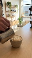 Barista pouring steamed milk into espresso cup creating latte art in modern cafe workspace with wooden desk, potted plants, and natural light from windows.