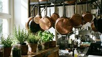 Rustic kitchen interior with copper pots and pans hanging from rack, fresh herbs in terracotta pots on wooden counter, warm natural lighting from window.