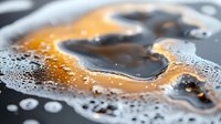 Macro shot of dark liquid with orange swirls and bubbles creating abstract patterns on reflective surface. Artistic close-up shows intricate details of fluid dynamics.