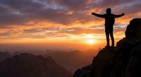 Male hiker silhouette with arms raised celebrating success on mountain peak at golden sunrise with dramatic orange sky and valley views below.