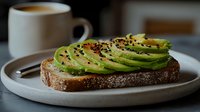 Fresh sliced avocado arranged on toasted sourdough bread with black sesame seeds and seasonings, served on ceramic plate with coffee cup in background.