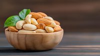 Mixed nuts and almonds in wooden bowl with fresh green leaves on rustic table for healthy snacking and cooking ingredients.