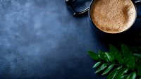 Hot chocolate drink with creamy foam in dark ceramic mug on moody blue concrete background with green tropical leaves and reading glasses, copy space.