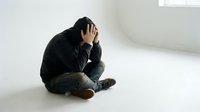 Depressed young man sitting on floor holding head in hands showing mental health struggles and emotional distress in minimalist white room setting.
