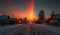 Winter village street at sunset with dramatic orange sky and snow-covered road leading through rural residential area with wooden houses and bare trees.