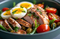 Grilled chicken breast slices with boiled eggs, cherry tomatoes, green beans and olives in ceramic bowl. Fresh herbs and spices garnish protein-rich meal.