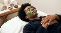 Young African American man relaxing with green facial mask at spa. Skincare treatment for men. Wellness and self-care routine in professional salon setting.