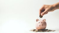 Hand dropping coin into pink piggy bank surrounded by scattered coins on white background for savings and financial planning concepts.