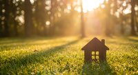 Small wooden house model on green grass with warm sunlight and bokeh background representing home ownership dreams and real estate investment concepts.