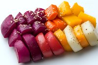Colorful rainbow gradient of sliced root vegetables arranged in rows, seasoned with sesame seeds and spices on white background, fresh healthy food preparation.
