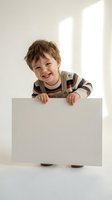 Happy toddler boy with curly brown hair smiling while holding blank white sign in bright room. Child wearing striped sweater and overalls for advertising mockup.