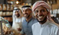 Middle Eastern man in traditional white thobe and red keffiyeh smiling warmly at cafe counter, with friends in background creating welcoming atmosphere.