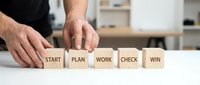 Male professional arranging wooden blocks spelling START PLAN WORK CHECK WIN on white desk in modern office environment for business strategy concepts.