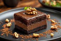 Rich chocolate mousse layered between dark chocolate cake, topped with ganache and walnuts on black plate, scattered cocoa powder and nuts create elegant dessert.