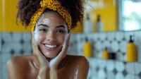 Young woman with dark curly hair and medium skin tone wearing golden headwrap, smiling at camera with hands on face in modern bathroom with yellow accents. Skincare and beauty routine concept.