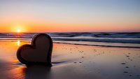 Wooden heart shape on wet sand beach at golden sunset with ocean waves and footprints creating romantic seaside scene for love and relationship concepts.