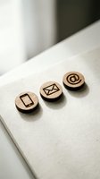 Three wooden circular tokens with engraved contact icons on white paper surface. Symbols represent mobile phone, email envelope, and at sign for communication channels and digital contact methods.