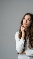 Young Caucasian woman in white shirt looking up thoughtfully with hand on chin, contemplating future plans and possibilities with copy space.