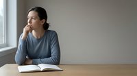 Thoughtful woman sitting at wooden table with open book, gazing out window in contemplative mood. Natural lighting creates peaceful atmosphere for reading.